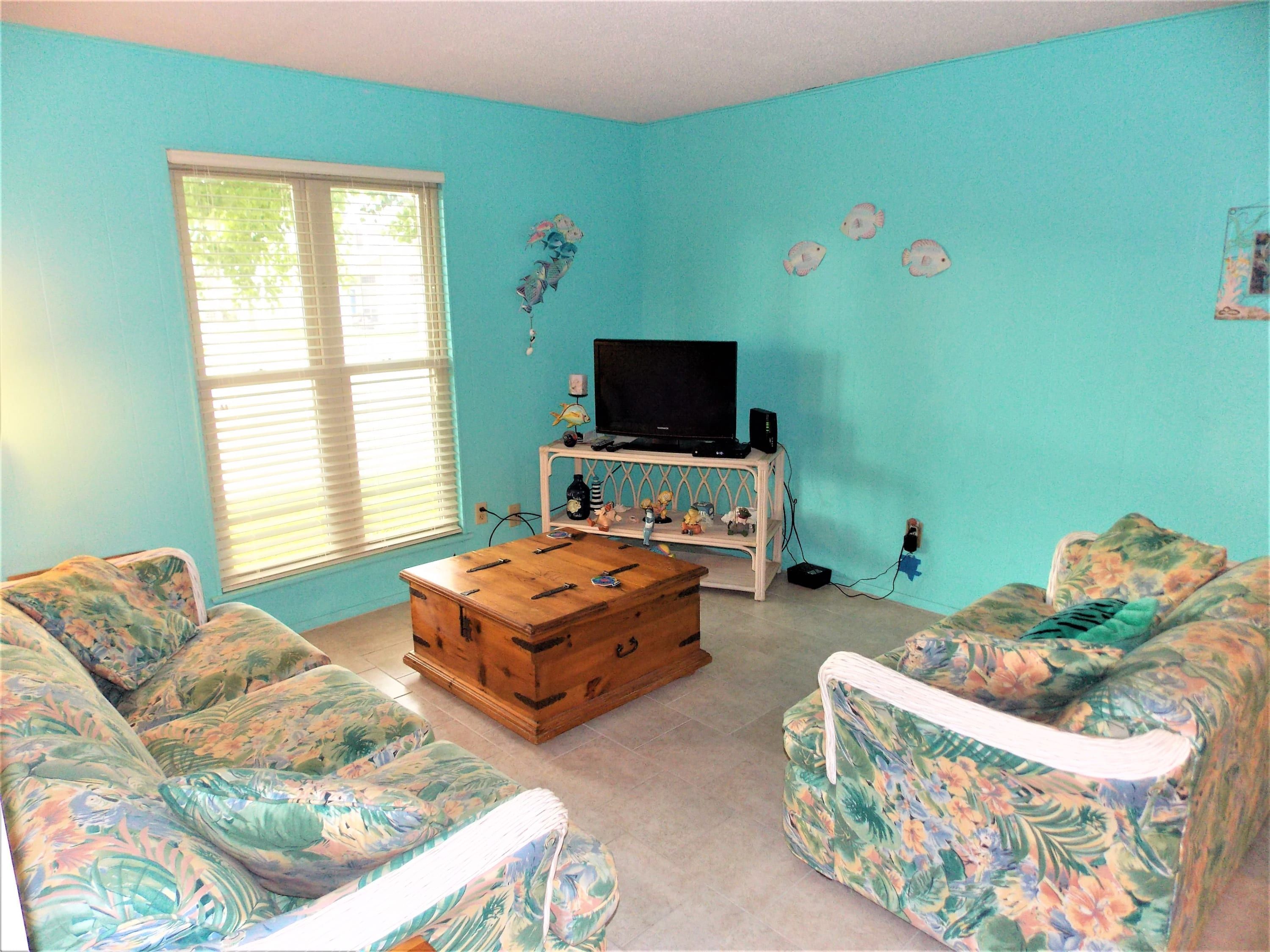 Une pièce aux murs de couleur turquoise, avec une table basse en bois, des canapés à motifs floraux, un téléviseur posé sur un meuble blanc et une fenêtre équipée de stores.