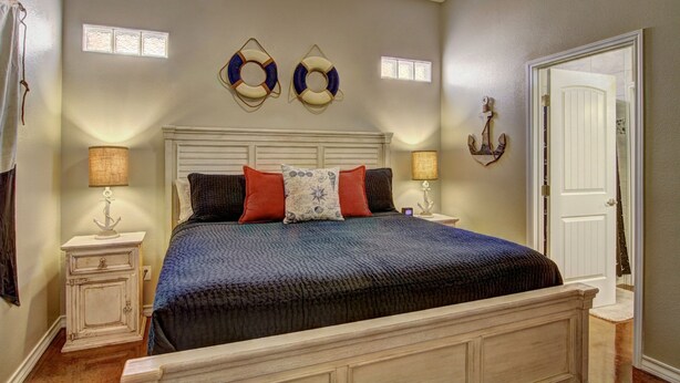 A bedroom with a bed, two nightstands, a door, and decorative wall items. - Happy Harbor 2 Resort Style Pools Playground (Port Aransas)