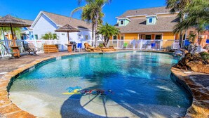 A swimming pool with a stone border, surrounded by houses, palm trees, and a wooden deck with chairs. - Happy Harbor 2 Resort Style Pools Playground (Port Aransas)