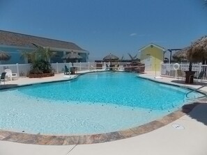 A clear swimming pool with a white fence surrounding it, a thatched-roof structure, and a yellow building in the background. - Happy Harbor 2 Resort Style Pools Playground (Port Aransas)