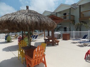 A beachside patio with a thatched umbrella, colorful Adirondack chairs, and a pool area. - Happy Harbor 2 Resort Style Pools Playground (Port Aransas)