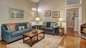 A living room with a blue sofa set, two side tables with lamps, a wooden coffee table, and a bookshelf. - Happy Harbor 2 Resort Style Pools Playground (Port Aransas)