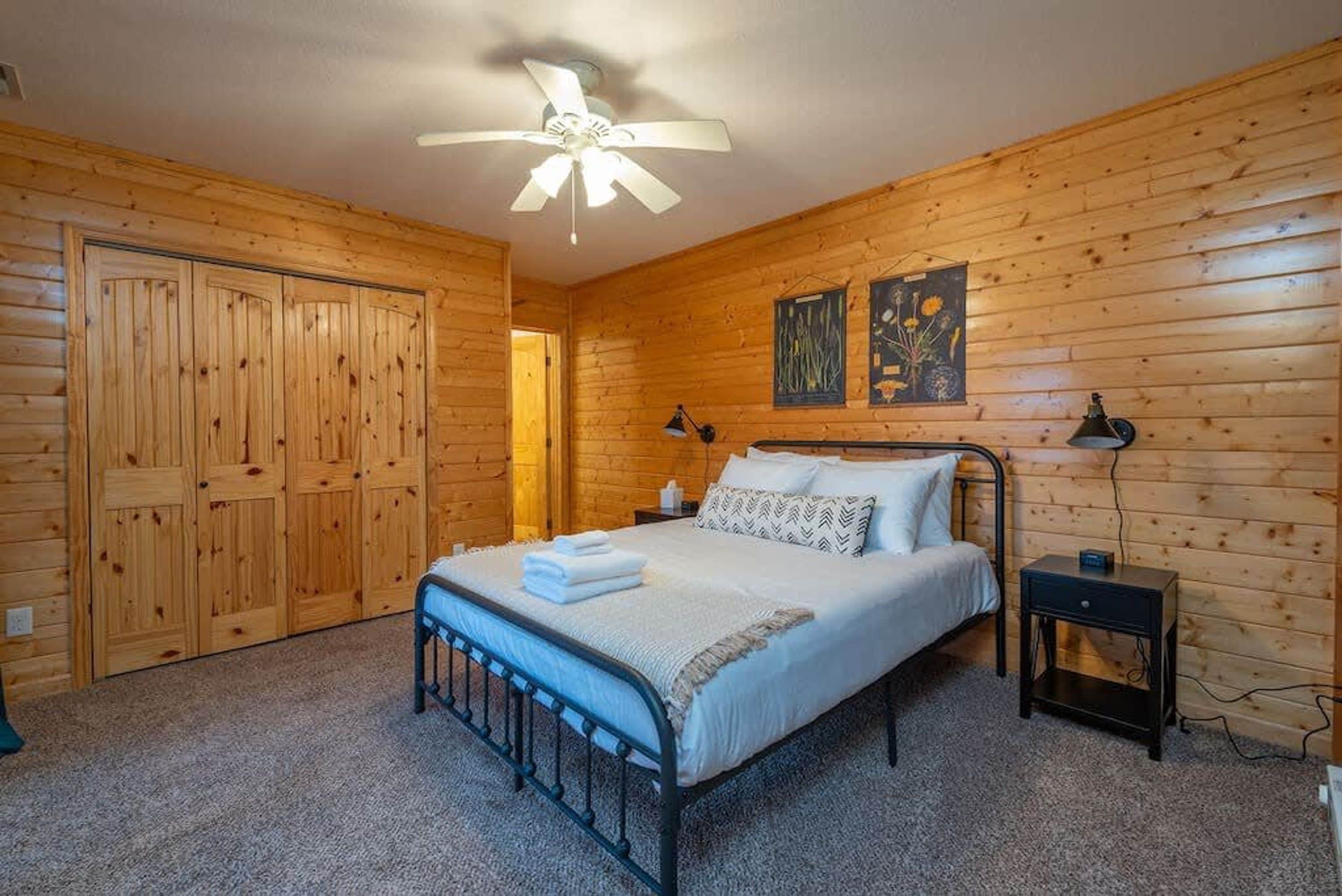 A bedroom with a bed, a nightstand, a ceiling fan, and wooden walls.