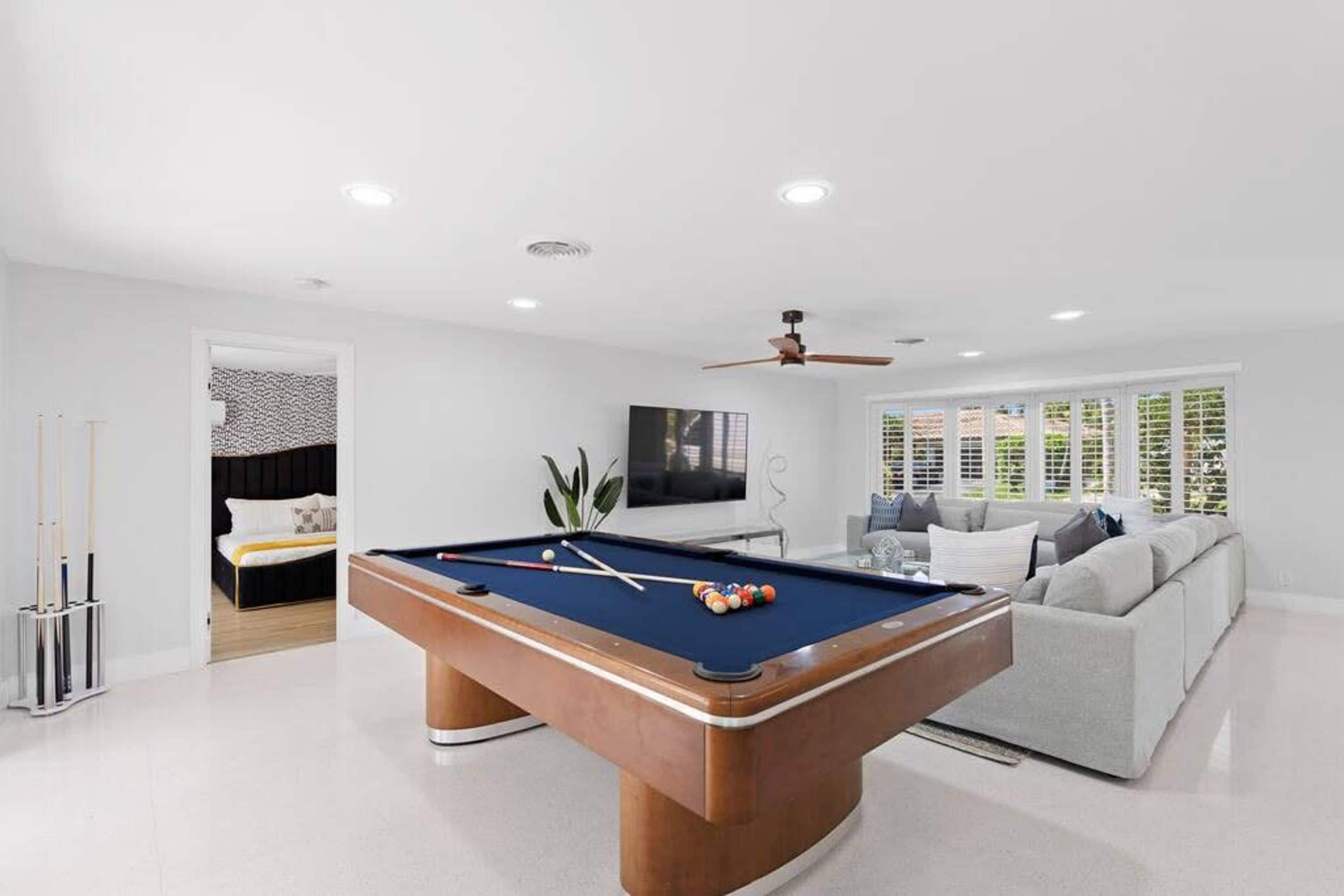 A pool table with balls, a sofa, and a ceiling fan in a room with white walls and a large window.