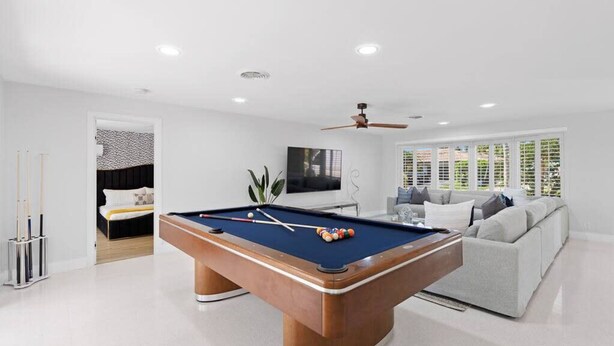 A pool table with balls, a sofa, and a ceiling fan in a room with white walls and a large window. - Heated Pool - Luxury Kitchen - 1 Mile to Beach (Fort Lauderdale)