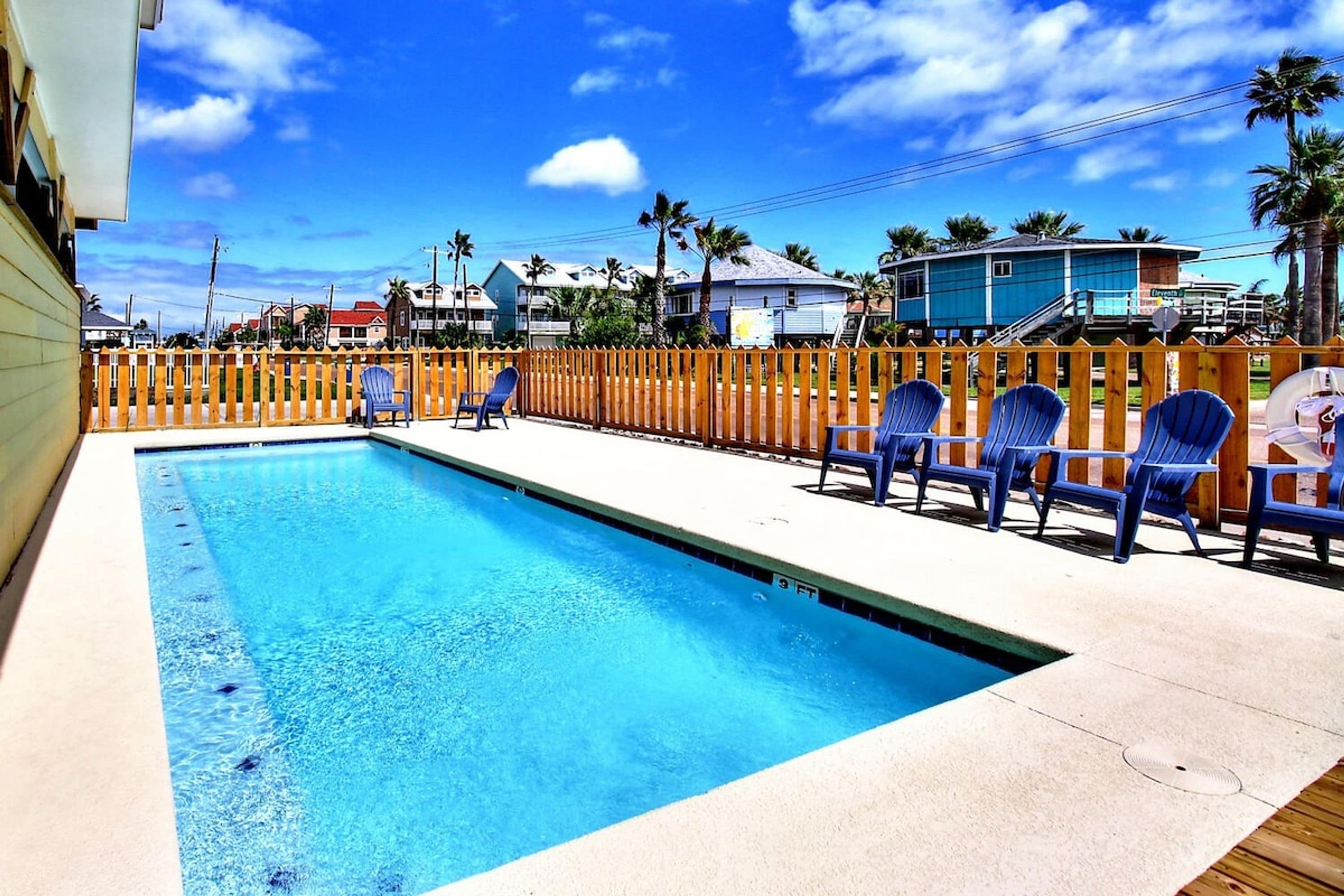 A swimming pool with a wooden deck and chairs, surrounded by a wooden fence.