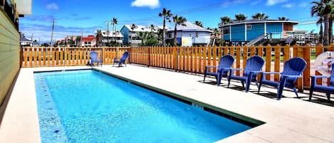 A swimming pool with a wooden deck and chairs, surrounded by a wooden fence.