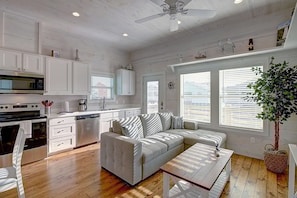 A modern kitchen with white cabinetry, a built-in oven, and a microwave. There is a dining area with a wooden table and a sofa. - Sea Clusion- Golf Cart Friendly Pet Friendly (Port Aransas)
