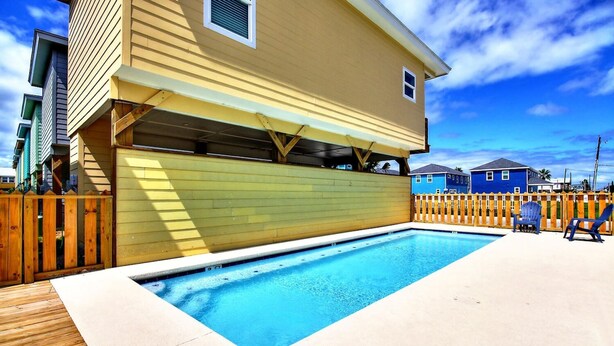 Una piscina residencial con muelle de madera, área cercada y una casa de dos pisos con techo plano.