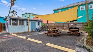 Zona residencial con una casa azul, un cobertizo pequeño y una valla de madera. Hay mesas de picnic y bicicletas aparcadas.