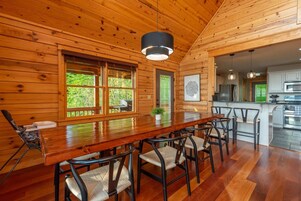A wooden interior with a dining area featuring a long table and chairs, large windows, and a kitchen area with modern appliances.
