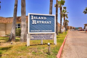 A sign for Island Retreat, indicating it is a beachfront condominium with a clear sky and palm trees in the background. - Island Oasis- Beach Access Pools Volleyball (Port Aransas)