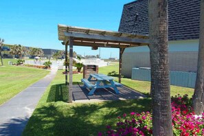 A covered outdoor seating area with a picnic table, surrounded by a well-maintained lawn and a pathway leading to a building. - Island Oasis- Beach Access Pools Volleyball (Port Aransas)