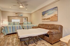 A hotel room with a bed, a sofa, a ceiling fan, and a painting on the wall. - Island Oasis- Beach Access Pools Volleyball (Port Aransas)