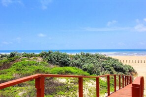 A wooden boardwalk leading to a beach with dense vegetation in the foreground and a clear view of the ocean. - Island Oasis- Beach Access Pools Volleyball (Port Aransas)