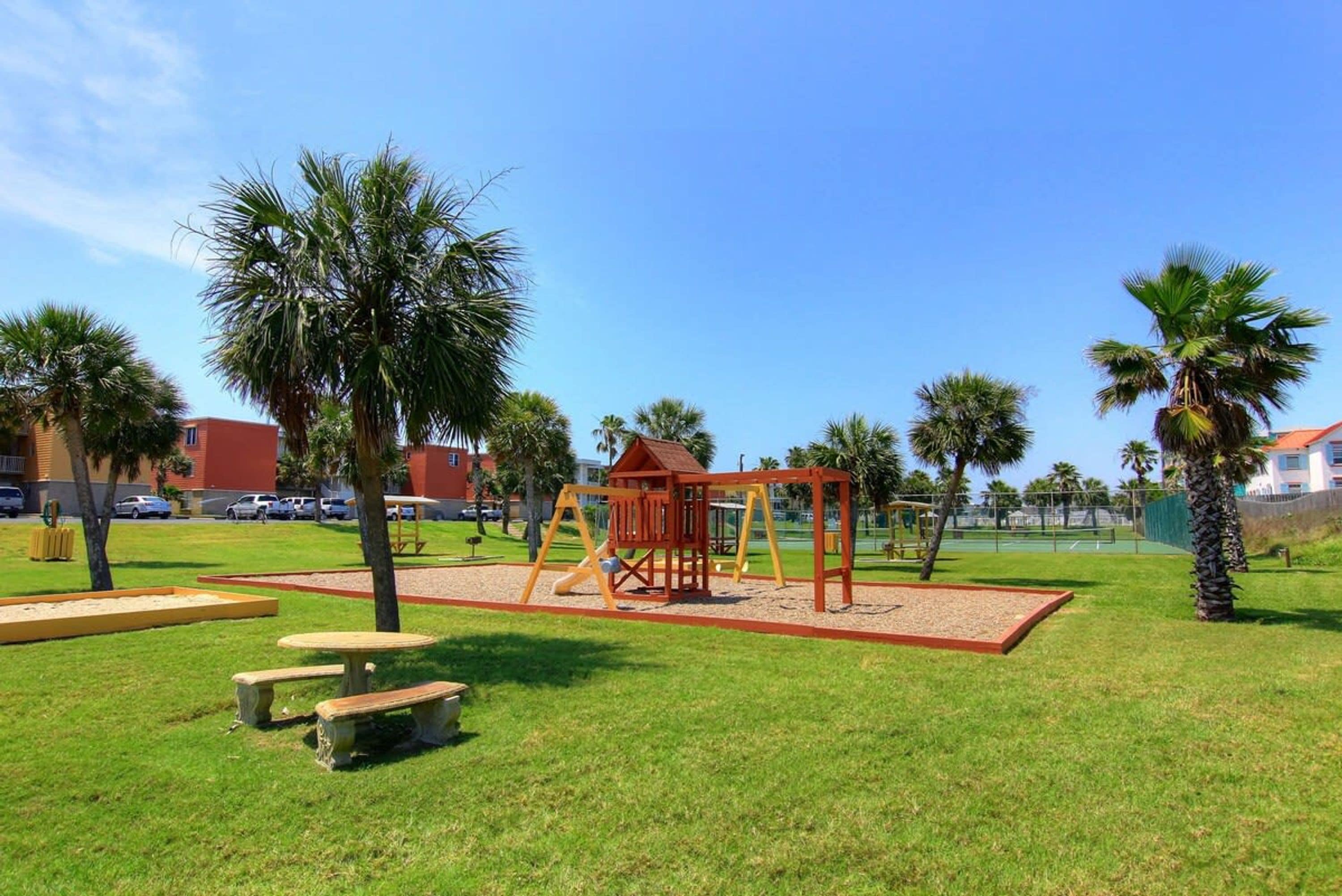A playground with a wooden swing set and a slide, surrounded by palm trees and a clear sky.
