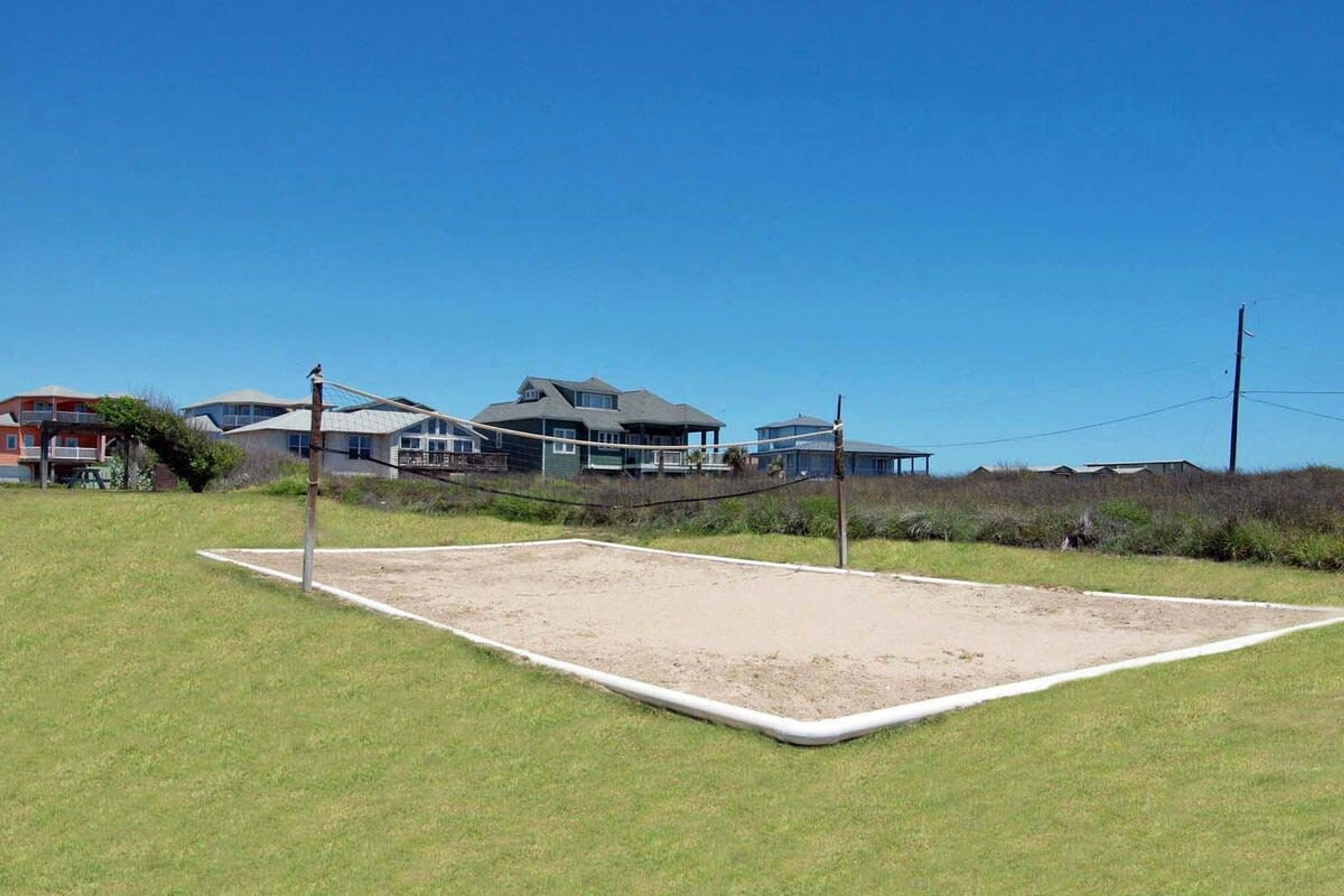 A volleyball court with a net, surrounded by grass and a few buildings in the background.
