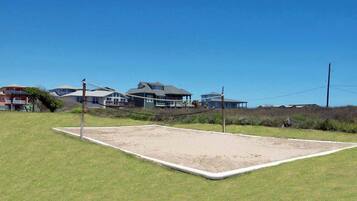 A volleyball court with a net, surrounded by grass and a few buildings in the background.