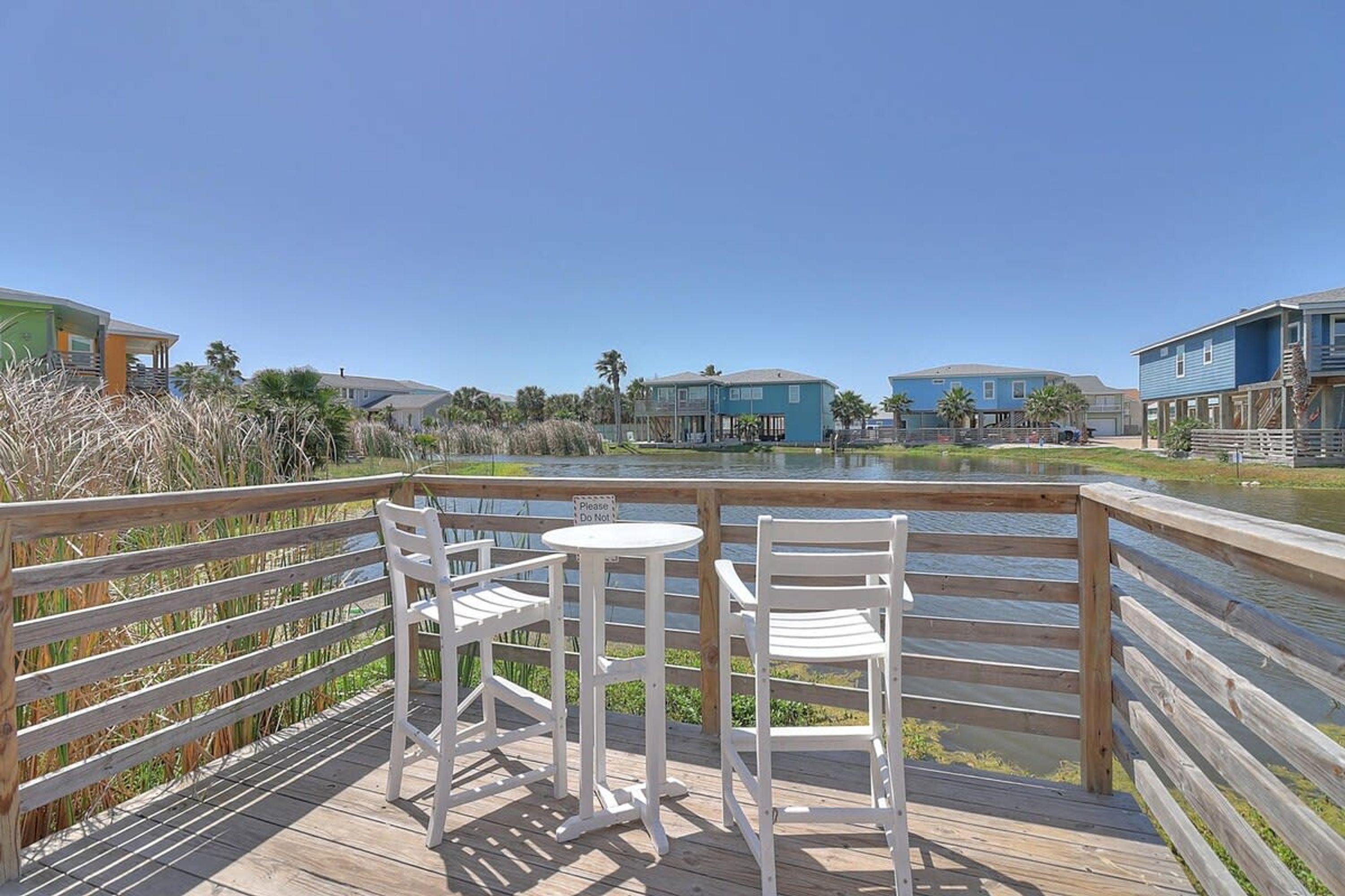 A wooden deck with two white chairs and a small table, overlooking a body of water with buildings in the background.