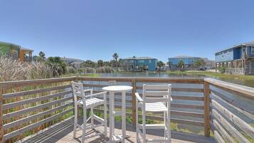 A wooden deck with two white chairs and a small table, overlooking a body of water with buildings in the background.