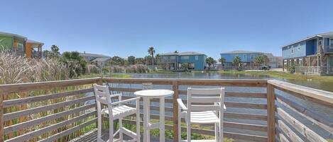A wooden deck with two white chairs and a small table, overlooking a body of water with buildings in the background.
