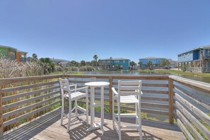 Um deck de madeira com duas cadeiras brancas e uma pequena mesa, com vista para um corpo d'água e edifícios ao fundo.