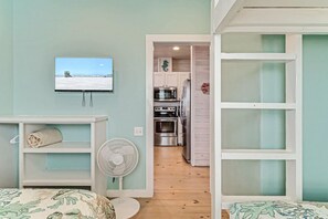 Een kamer met een televisie waarop een strandafbeelding wordt weergegeven, een ventilator en een kitchenette met een magnetron en oven.