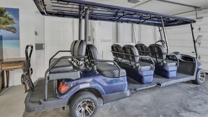 Um carrinho de golfe azul com teto, estacionado em uma garagem com porta branca e um quadro de uma paisagem praiana.