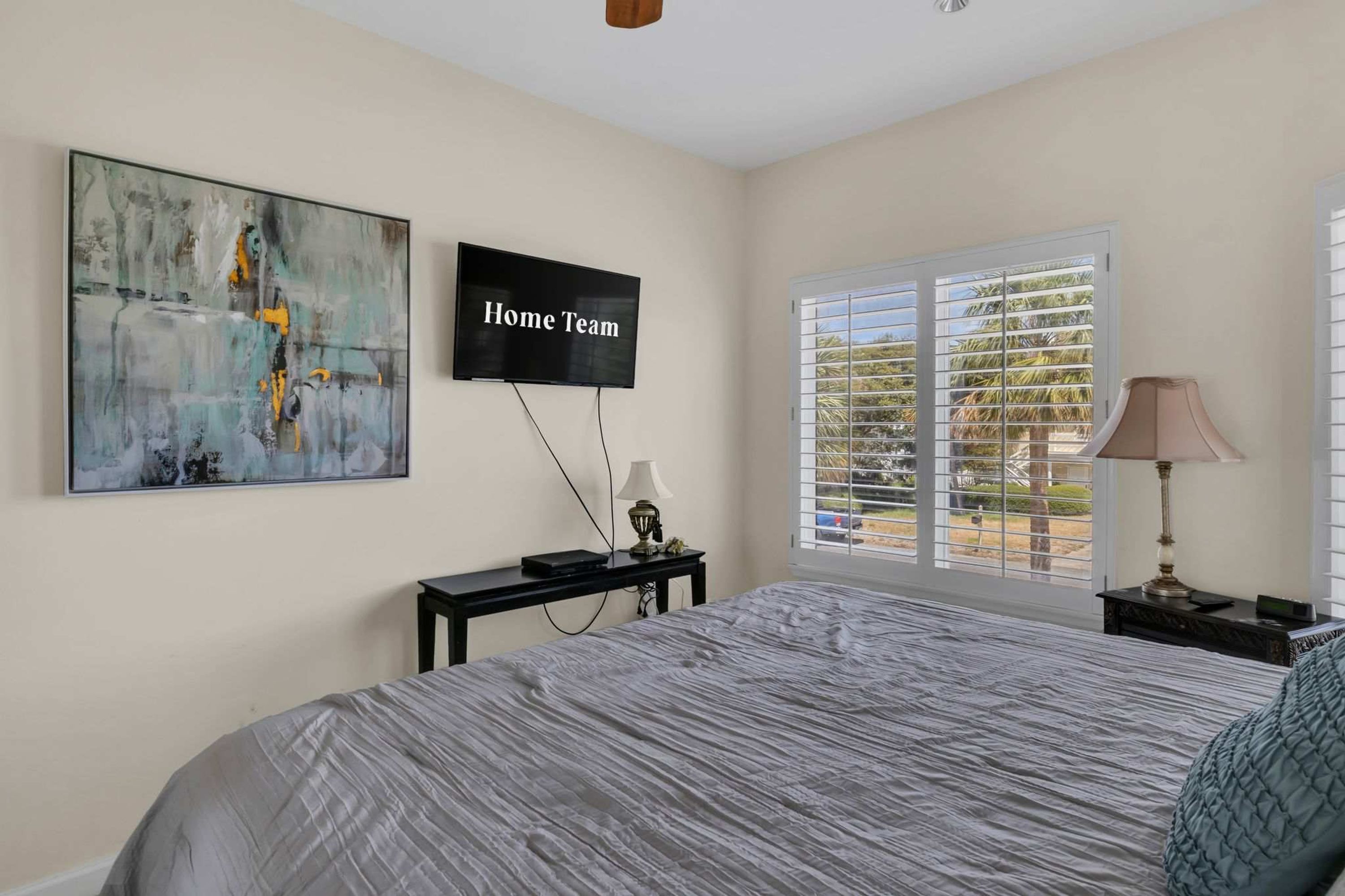 A bedroom with a bed, a TV, a desk, and a window with blinds.