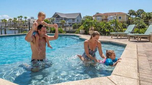 Uma piscina com uma família aproveitando a água, cercada por cadeiras de descanso e um céu claro.