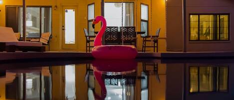 A swimming pool with a flamingo float, surrounded by a covered patio with string lights.