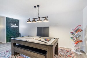 A pool table with a neon sign, a bookshelf with board games, and a billiards cue rack.