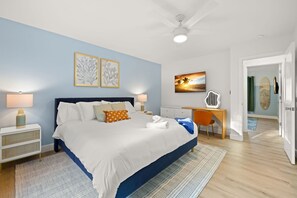 Ein Schlafzimmer mit Bett, Nachttischen, Schreibtisch, Stuhl, Deckenventilator, Kunstwerken und Blick auf den Strand durch das Fenster.