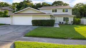 A two-story residential house with a garage, a well-maintained lawn, and a clear sky. - Hot Tub Sportscourt Game Room 5 min to Beach (Seminole)