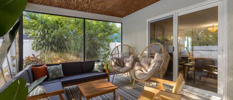 Une terrasse ensoleillée avec un plafond en bois, un canapé, deux fauteuils en osier, une table à manger et une vue sur la verdure environnante.
