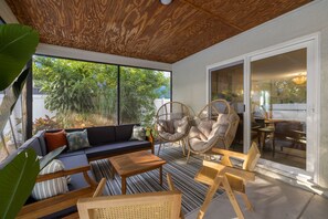 Um pátio ensolarado com teto de madeira, um sofá, duas cadeiras de vime, uma mesa de jantar e vista para o verde lá fora.