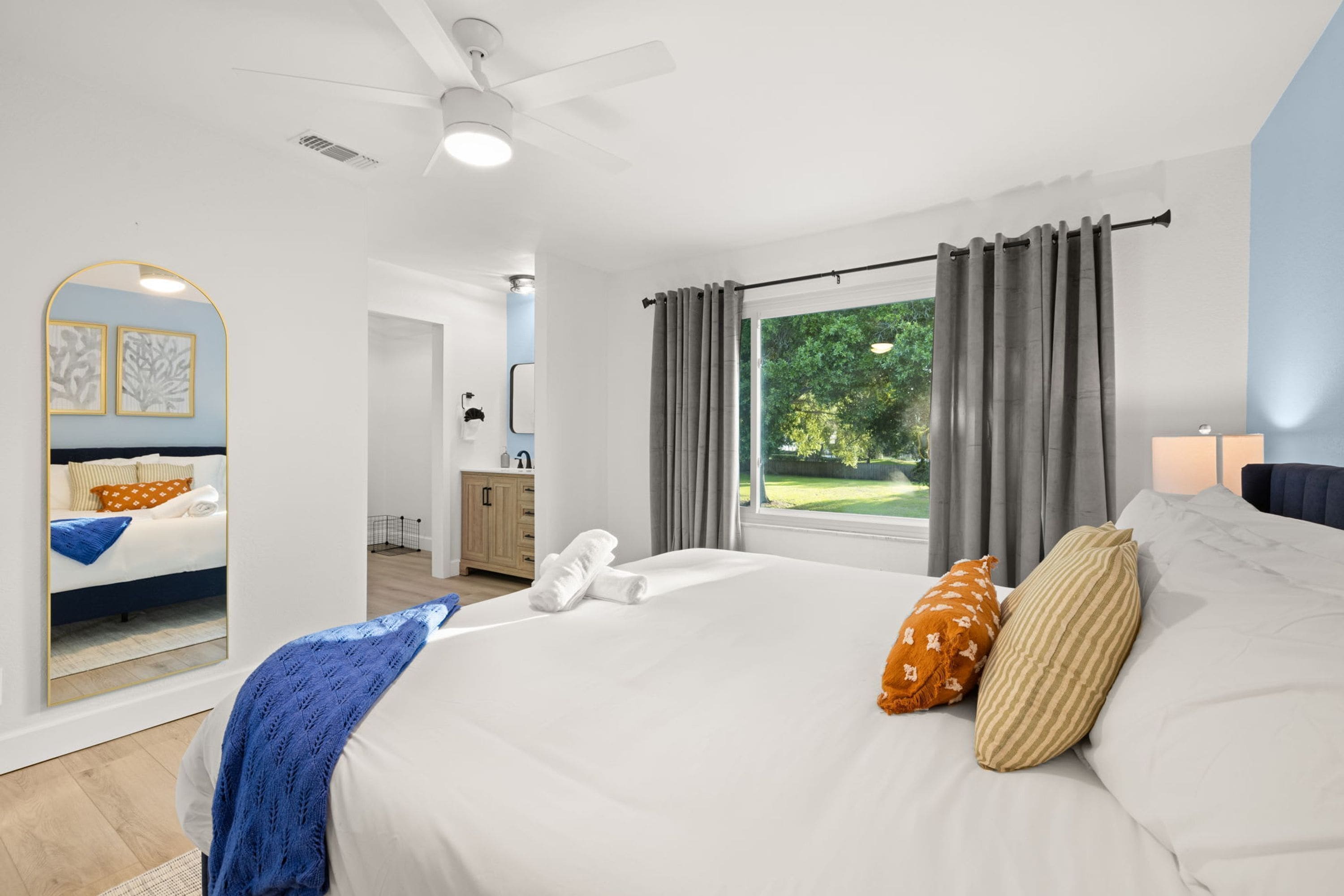 A modern bedroom with a large bed, a ceiling fan, a window with curtains, and a view of greenery outside.