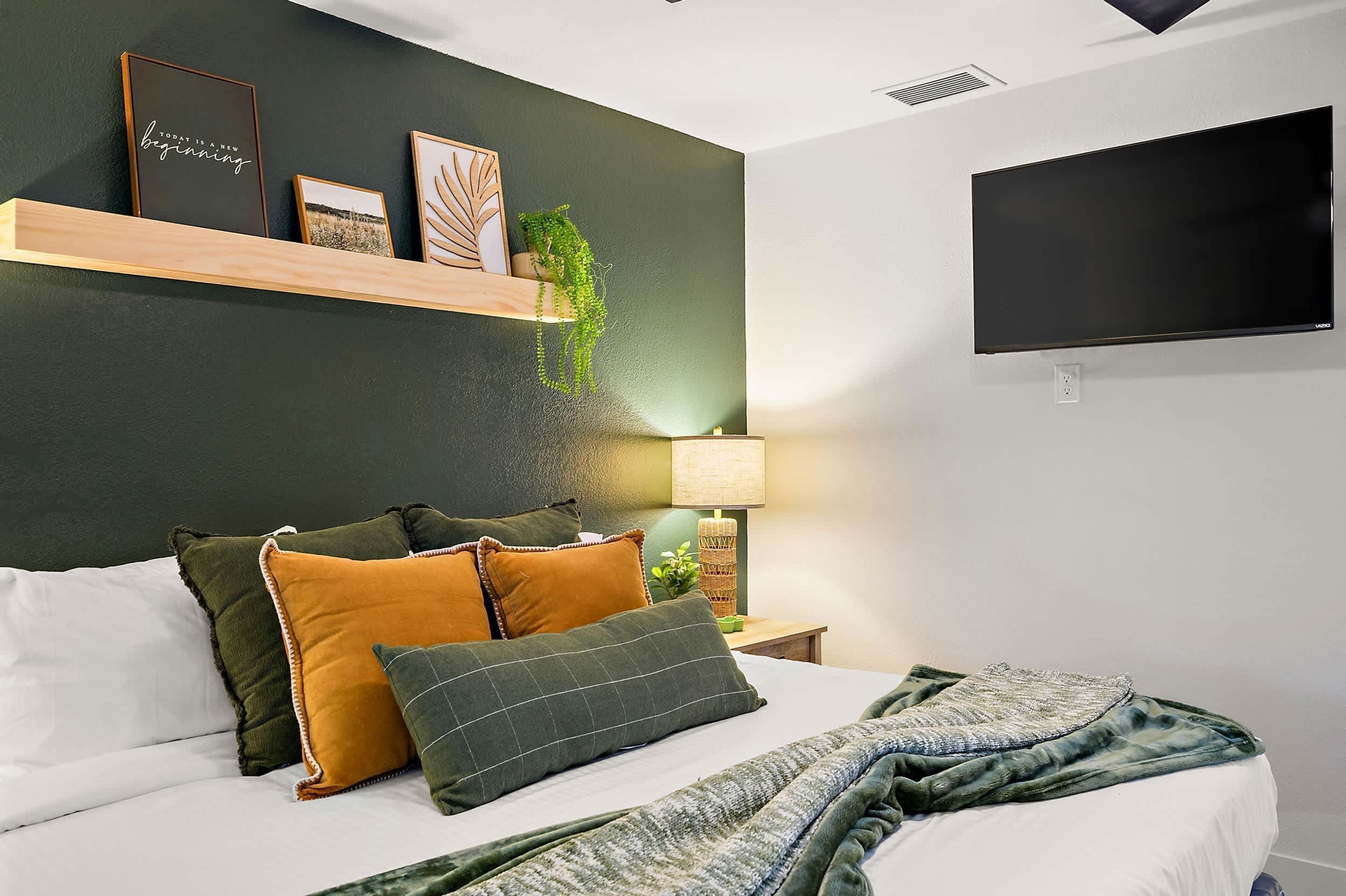 Un dormitorio con cama, almohadas, una mesita de noche con lámpara, un televisor montado en la pared y un estante con objetos decorativos.