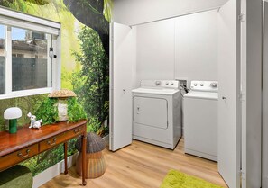Ein kleiner Hauswirtschaftsraum mit Waschmaschine und Trockner, ein Holztisch mit Lampe sowie eine Wand mit einem Wandgemälde von üppigem Grünwerk.