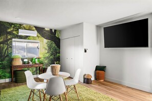A modern living room with a dining area, a large window, and a wall-mounted TV. - The Desert Emerald Pool Hot Tub Games More (Scottsdale)