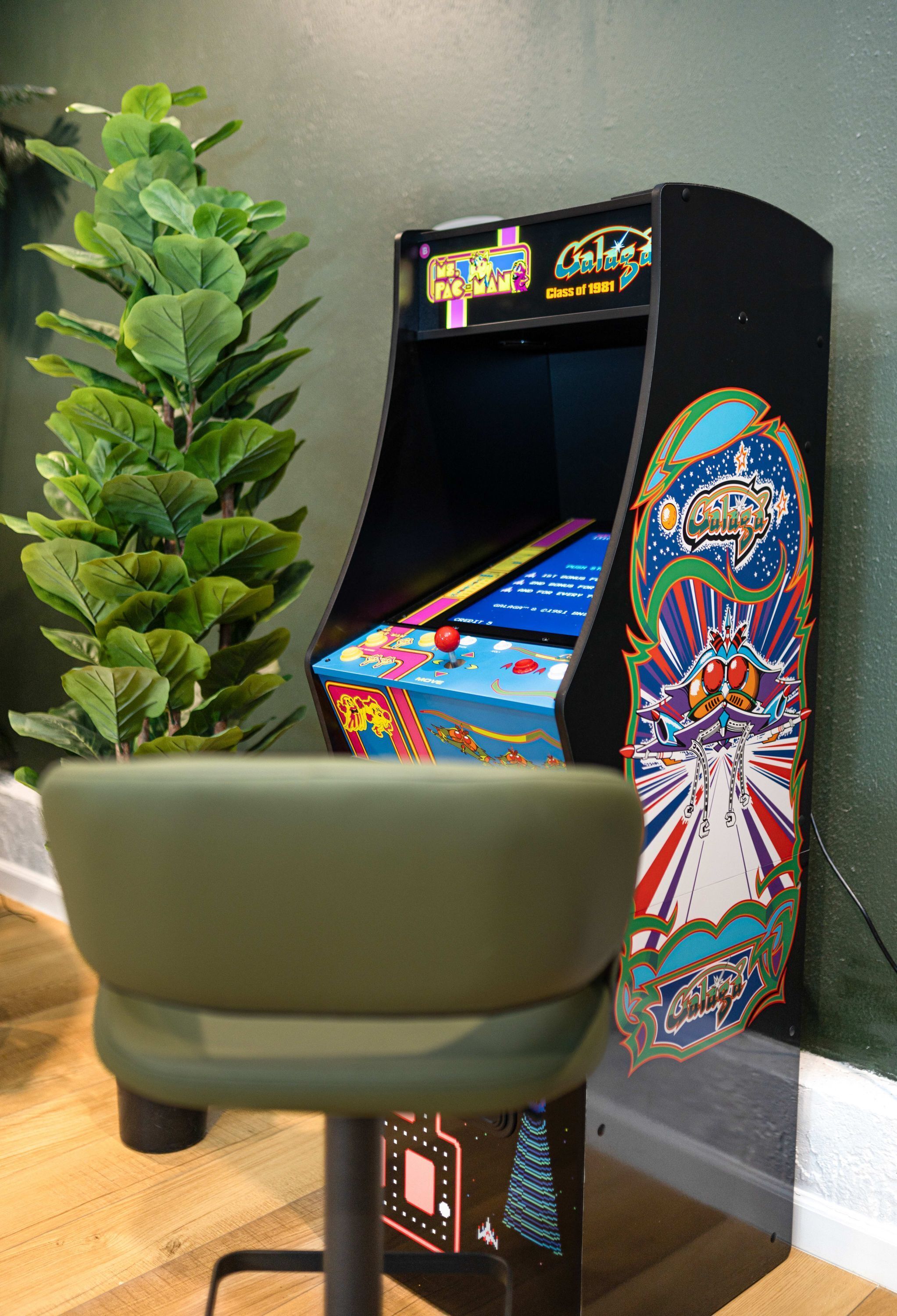 Une borne d’arcade ancienne, dotée d’un écran aux couleurs vives, avec une chaise verte devant et une plante en pot à côté.