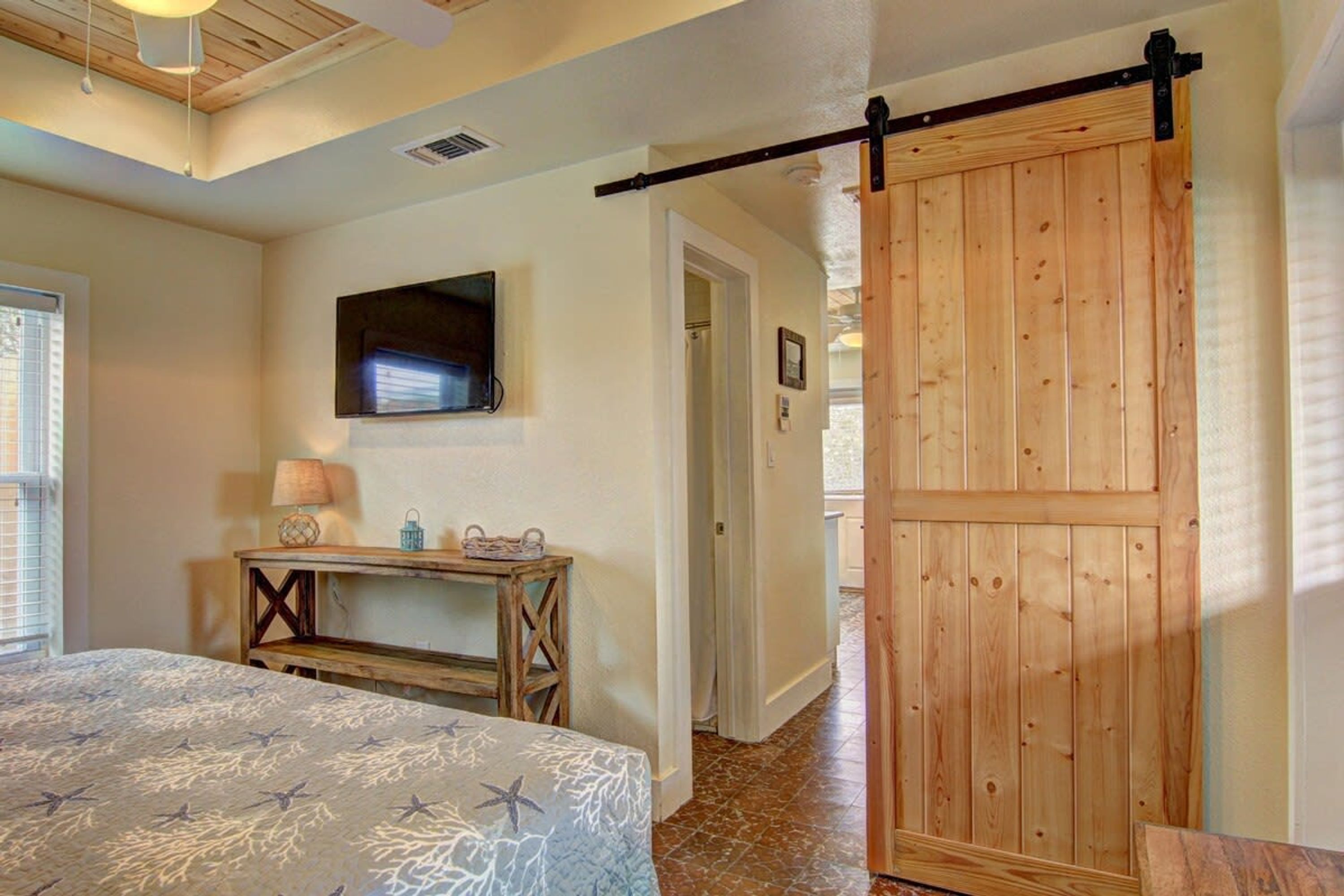 Un dormitorio con cama, televisor montado en la pared, puerta de madera y un pasillo que conduce a otra habitación.