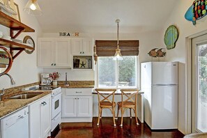 Uma cozinha com armários brancos, uma mesa de jantar de madeira com duas cadeiras, uma geladeira e uma janela com persianas.