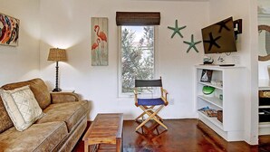 Ein Wohnzimmer mit braunem Sofa, einem Holztisch, einem Regiestuhl, einem Fernseher auf einem Schrank und einem Fenster mit Blick auf Bäume.