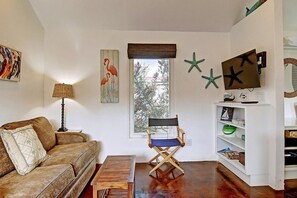 A living room with a brown sofa, a wooden table, a director's chair, a television on a stand, and a window with a view of trees. - Cozy Private Cottage Pet Friendly Prime Location (Port Aransas)