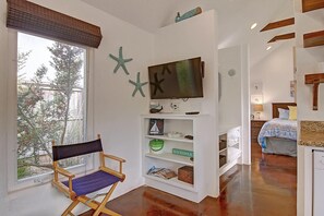 A room with a large window, a flat-screen TV mounted on the wall, a wooden chair, and a bookshelf with various items. - Cozy Private Cottage Pet Friendly Prime Location (Port Aransas)