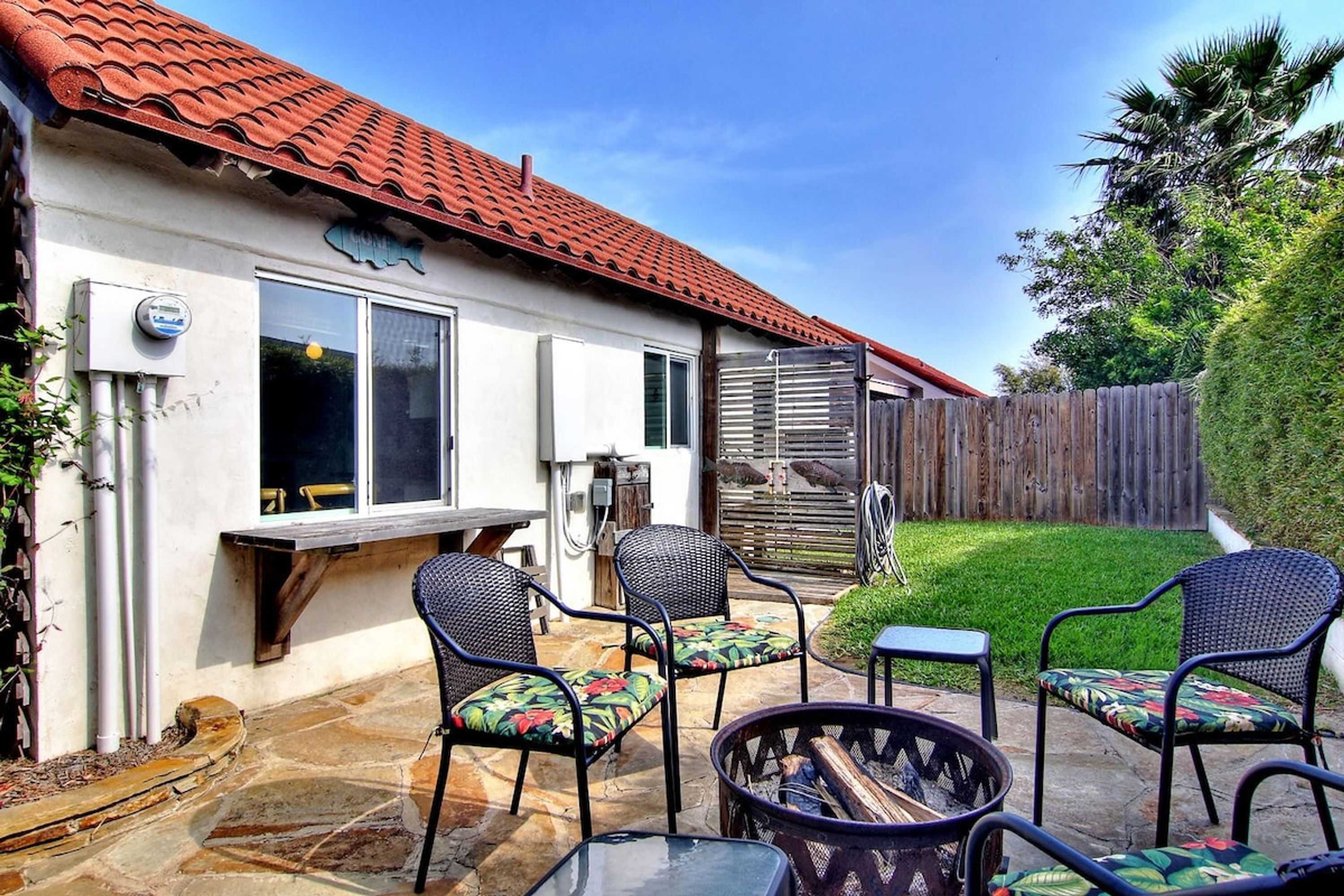 Uma casa com telhado de telhas vermelhas, um pátio com móveis de exterior e uma cerca de madeira ao fundo.