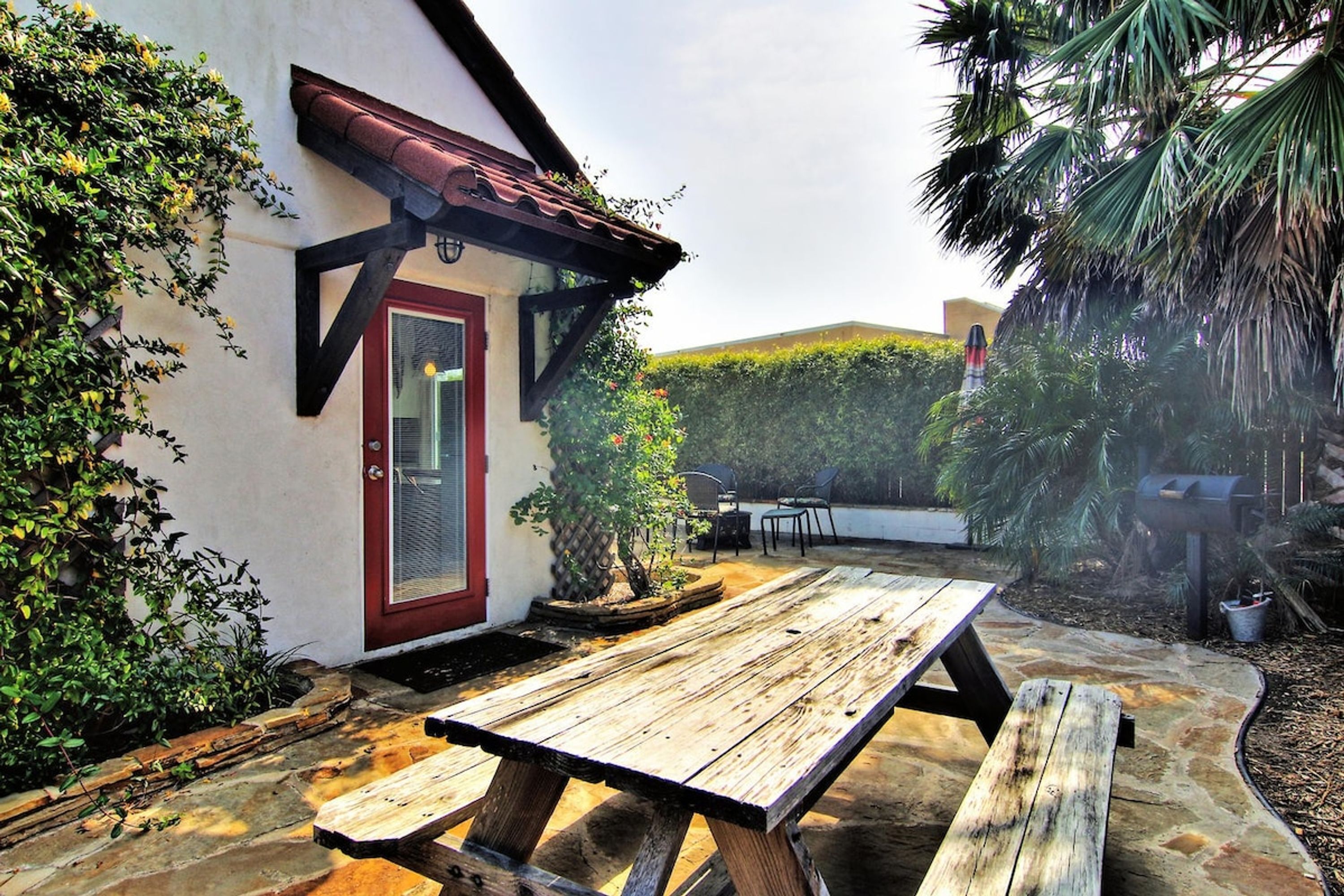 Una mesa de picnic de madera en un jardín, con un edificio blanco, una puerta roja y una valla metálica.
