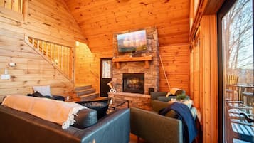 A log cabin interior with a stone fireplace, a television mounted above it, and a staircase leading to an upper level.