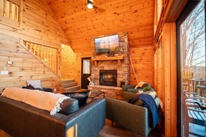 A log cabin interior with a stone fireplace, a television mounted above it, and a staircase leading to an upper level. - The Triple Timber in Mars Hill (Mars Hill)